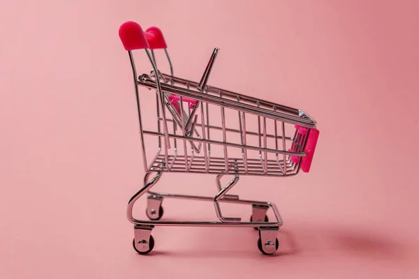 Small supermarket grocery push cart for shopping toy with wheels isolated on pink pastel colorful paper trendy background. Sale buy mall market shop consumer concept. Copy space