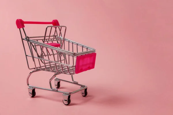 Small supermarket grocery push cart for shopping toy with wheels isolated on pink pastel colorful paper trendy background. Sale buy mall market shop consumer concept. Copy space