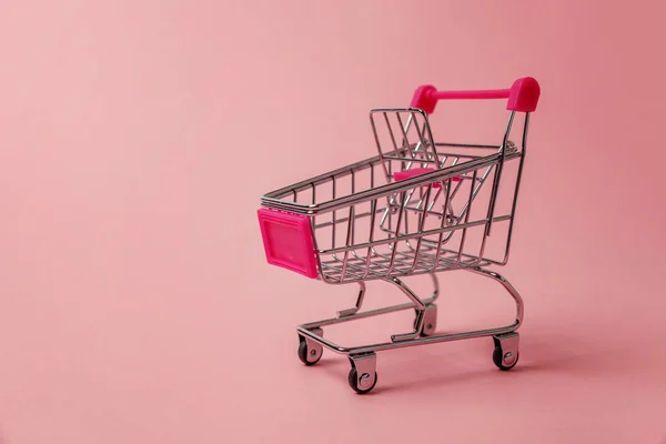 Small supermarket grocery push cart for shopping toy with wheels isolated on pink pastel colorful paper trendy background. Sale buy mall market shop consumer concept. Copy space