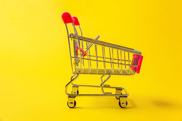 Small supermarket grocery push cart for shopping toy with wheels isolated on yellow colourful trendy modern fashion background. Sale buy mall market shop consumer concept. Copy space