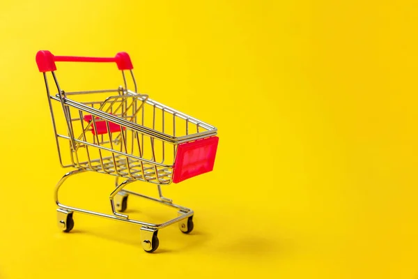 Small supermarket grocery push cart for shopping toy with wheels isolated on yellow colourful trendy modern fashion background. Sale buy mall market shop consumer concept. Copy space