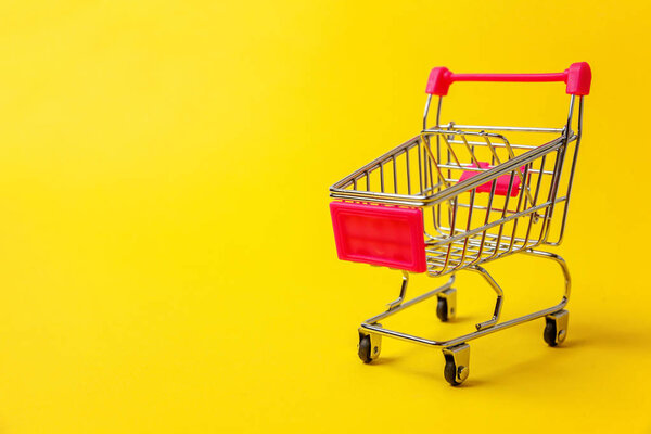 Small supermarket grocery push cart for shopping toy with wheels isolated on yellow colourful trendy modern fashion background. Sale buy mall market shop consumer concept. Copy space