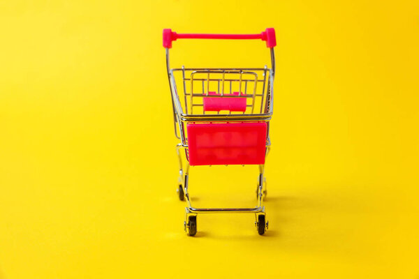 Small supermarket grocery push cart for shopping toy with wheels isolated on yellow colourful trendy modern fashion background. Sale buy mall market shop consumer concept. Copy space