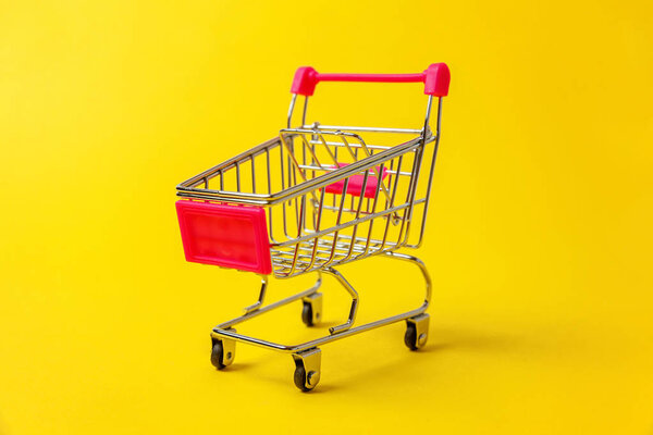 Small supermarket grocery push cart for shopping toy with wheels isolated on yellow colourful trendy modern fashion background. Sale buy mall market shop consumer concept. Copy space