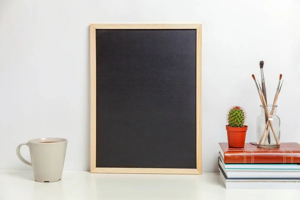 Home or office decor with mock up blank chalkboard on table near white wall. Minimalism interior workplace artist supplies decoration background. Hygge scandinavian style workspace concept