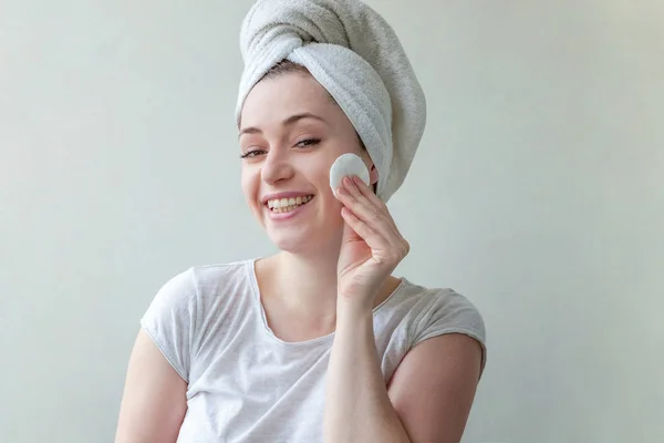 Beauty portrait of a smiling brunette woman with towel on head with soft healthy skin removing make up with cotton pad isolated over white background. Skincare cleansing spa relax concept