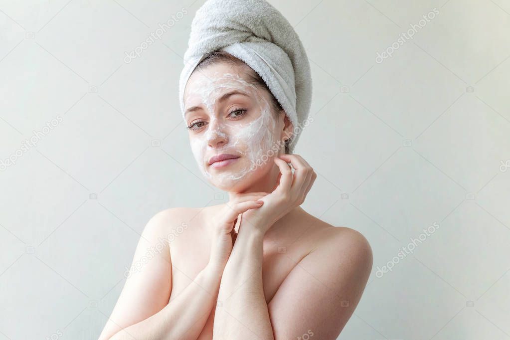 Beauty portrait of a smiling brunette woman in a towel on the head applying white nourishing mask or creme on face on white background isolated. Skincare cleansing spa relax concept