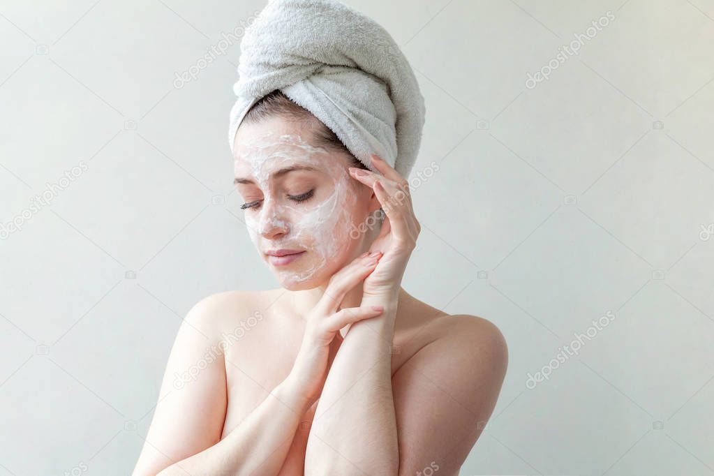 Beauty portrait of a smiling brunette woman in a towel on the head applying white nourishing mask or creme on face on white background isolated. Skincare cleansing spa relax concept