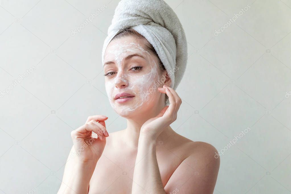 Beauty portrait of a smiling brunette woman in a towel on the head applying white nourishing mask or creme on face on white background isolated. Skincare cleansing spa relax concept