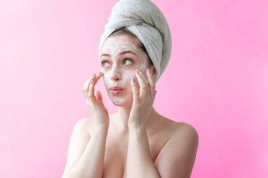 Bir havlu izole pembe arka plan yüzünde beyaz besleyici maske veya krem uygulayarak kafasına gülümseyen bir esmer kadın güzellik portresi. Cilt Bakımı temizlik spa kavramı relax