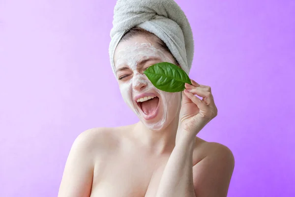 一个微笑的黑发女人的美丽的肖像在毛巾在头与白色营养面具或奶油在面孔和绿叶在手在紫色背景隔绝 护肤清洁生态有机美容水疗放松的概念 — 图库照片