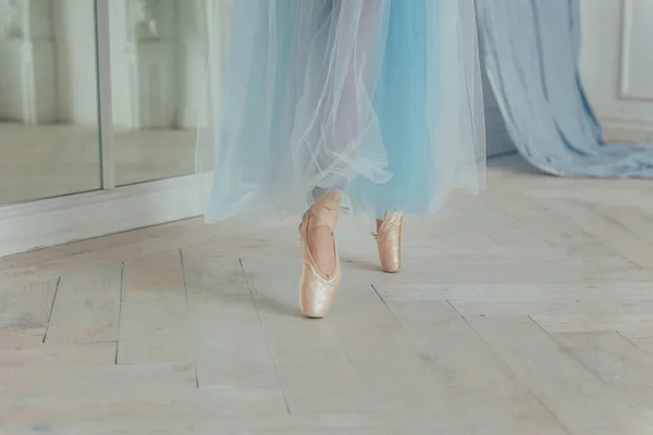 Hands of ballerina in blue tutu skirt puts on pointe shoes on leg in white light hall. Young classical ballet dancer woman in dance class