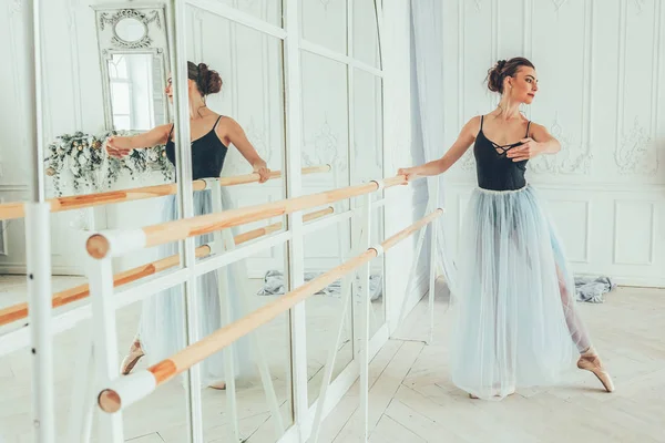 Young classical ballet dancer woman in dance class. Beautiful graceful ballerina practice ballet positions in blue tutu skirt near large mirror in white light hall