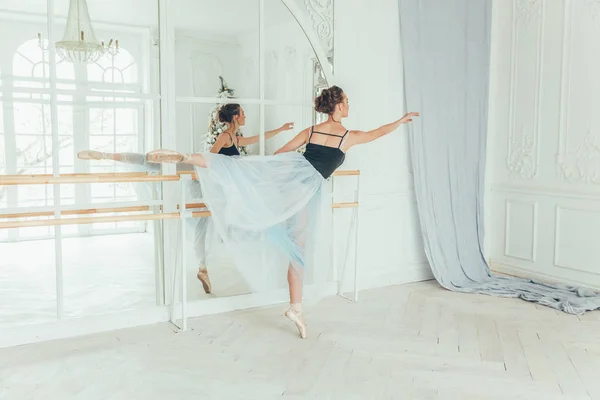 Young classical ballet dancer woman in dance class. Beautiful graceful ballerina practice ballet positions in blue tutu skirt near large mirror in white light hall