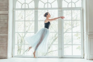 Genç Klasik Bale dansçısı kadın dans dersi. Mavi tutu etek beyaz ışık salonda büyük pencere yakınındaki güzel zarif balerin pratik bale pozisyonlar