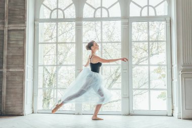Genç Klasik Bale dansçısı kadın dans dersi. Mavi tutu etek beyaz ışık salonda büyük pencere yakınındaki güzel zarif balerin pratik bale pozisyonlar