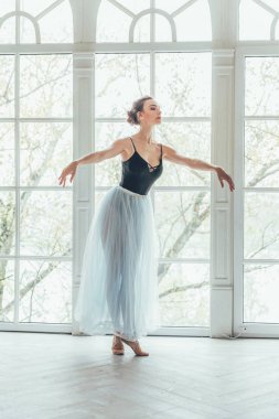 Genç Klasik Bale dansçısı kadın dans dersi. Mavi tutu etek beyaz ışık salonda büyük pencere yakınındaki güzel zarif balerin pratik bale pozisyonlar