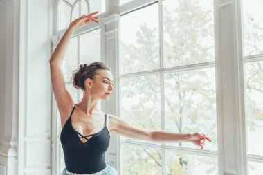 Genç Klasik Bale dansçısı kadın dans dersi. Mavi tutu etek beyaz ışık salonda büyük pencere yakınındaki güzel zarif balerin pratik bale pozisyonlar