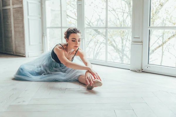 Young classical ballet dancer woman in dance class. Beautiful graceful ballerina practice ballet positions in blue tutu skirt near large window in white light hall
