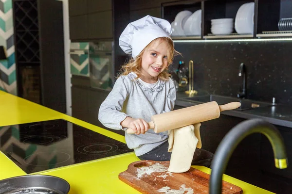 Little girl with chef hat, rolling pin preparing dough, bake homemade holiday apple pie in kitchen. Kid cooking healthy food at home and having fun. Childhood, household, teamwork helping concept