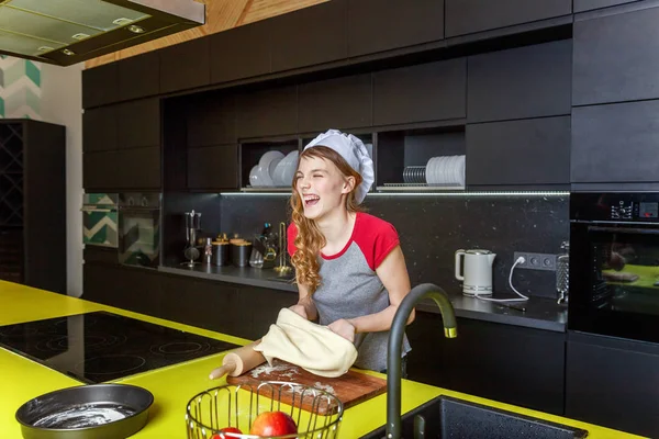 Teenage girl with chef hat, rolling pin preparing dough, bake homemade holiday apple pie in kitchen. Young woman cooking healthy food at home and having fun. Food, healthy eating, household concept