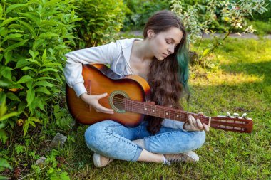 Young hipster woman sitting in grass and playing guitar on park or garden background. Teen girl learning to play song and writing music. Hobby, lifestyle, relax, Instrument, leisure, education concept clipart