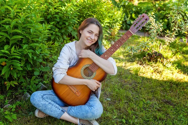 Young hipster woman sitting in grass and playing guitar on park or garden background. Teen girl learning to play song and writing music. Hobby, lifestyle, relax, Instrument, leisure, education concept