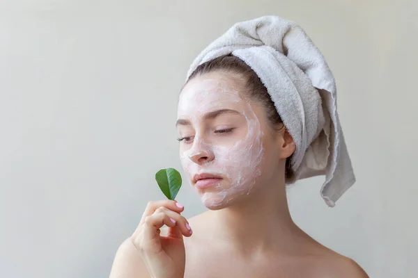 Beauty portrait of woman in towel on head with white nourishing mask or creme on face and green leaf in hand on white background isolated. Skincare cleansing eco organic cosmetic spa relax concept