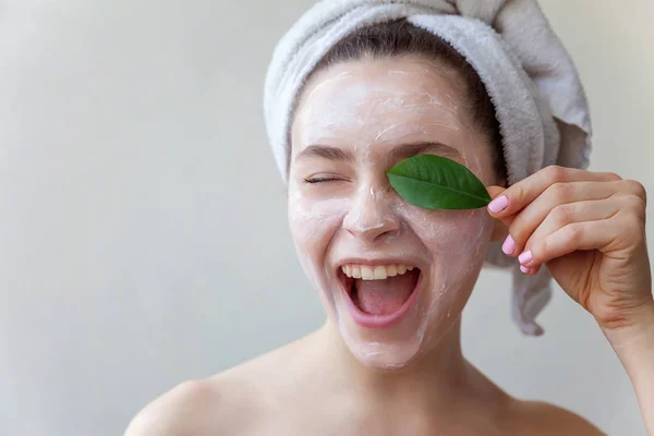 Beauty portrait of woman in towel on head with white nourishing mask or creme on face and green leaf in hand on white background isolated. Skincare cleansing eco organic cosmetic spa relax concept