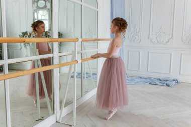 Dans sınıfındaki genç klasik balerin kız. Güzel, zarif balerin, bale pozisyonu pembe etekli, beyaz ışıklı salonda büyük aynanın yanında.