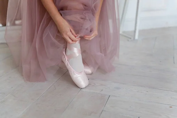 Hands of ballerina in pink tutu skirt puts on pointe shoes on leg in white light hall. Young classical ballet dancer girl in dance class