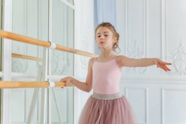 Dans sınıfındaki genç klasik balerin kız. Güzel, zarif balerin, bale pozisyonu pembe etekli, beyaz ışıklı salonda büyük aynanın yanında.