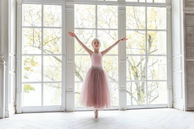 Dans sınıfındaki genç klasik balerin kız. Güzel, zarif balerin, bale pozisyonu pembe etekli, beyaz ışıklı salonun geniş penceresinin yanında.