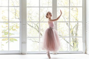Dans sınıfındaki genç klasik balerin kız. Güzel, zarif balerin, bale pozisyonu pembe etekli, beyaz ışıklı salonun geniş penceresinin yanında.