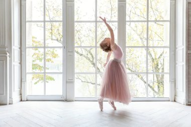 Dans sınıfındaki genç klasik balerin kız. Güzel, zarif balerin, bale pozisyonu pembe etekli, beyaz ışıklı salonun geniş penceresinin yanında.