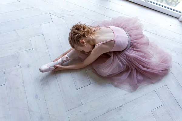 Young classical ballet dancer girl in dance class. Beautiful graceful ballerina in pink tutu skirt puts on pointe shoes near large window in white light hall