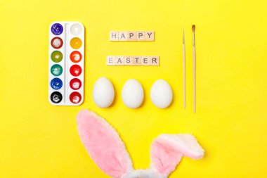 Mutlu Paskalya kavramı. Tatil için hazırlık. Yazıt Mutlu Paskalya yumurta renkli boyalar ve trendy sarı arka plan üzerinde izole tavşan kulakları mektuplar. Üstten görünüm kopya alanı basit minimalizm düz yatıyordu
