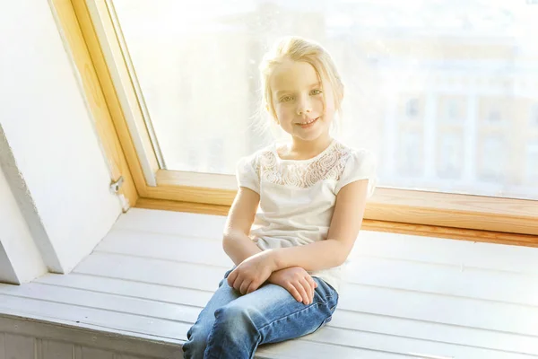 かわいい甘い笑顔女の子のジーンズと白 シャツに明るい光の居間で自宅の窓枠に座っていると子供のころ 青少年を考えてリラックス コンセプト — ストック写真