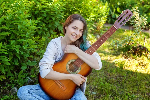 Young hipster woman sitting in grass and playing guitar on park or garden background. Teen girl learning to play song and writing music. Hobby, lifestyle, relax, Instrument, leisure, education concept