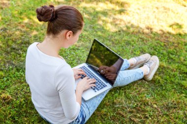 Serbest iş kavramı. Dizüstü pc bilgisayar üzerinde çalışan Şehir Parkı yeşil çim Çimenlikte oturan genç kadın. Yaşam tarzı otantik samimi öğrenci kız açık havada okuyor. Mobil ofis