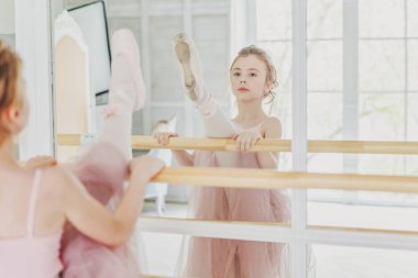 Dans sınıfındaki genç klasik balerin kız. Güzel, zarif balerin, bale pozisyonu pembe etekli, beyaz ışıklı salonda büyük aynanın yanında.
