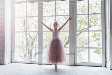 Dans sınıfındaki genç klasik balerin kız. Güzel, zarif balerin, bale pozisyonu pembe etekli, beyaz ışıklı salonun geniş penceresinin yanında.