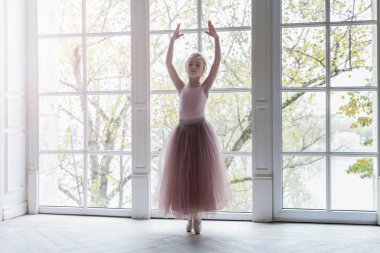 Dans sınıfındaki genç klasik balerin kız. Güzel, zarif balerin, bale pozisyonu pembe etekli, beyaz ışıklı salonun geniş penceresinin yanında.