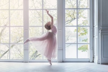 Dans sınıfındaki genç klasik balerin kız. Güzel, zarif balerin, bale pozisyonu pembe etekli, beyaz ışıklı salonun geniş penceresinin yanında.