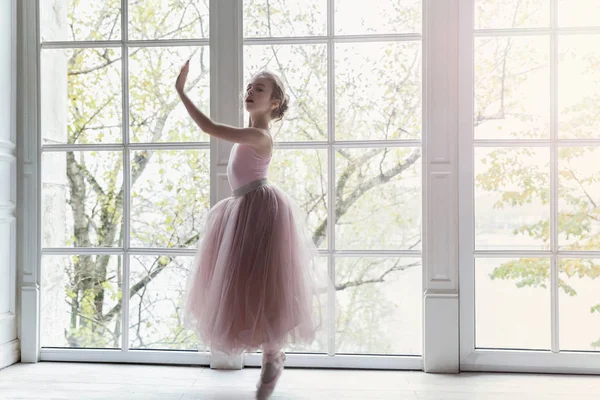 Young classical ballet dancer girl in dance class. Beautiful graceful ballerina practice ballet positions in pink tutu skirt near large window in white light hall
