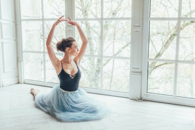 Genç Klasik Bale dansçısı kadın dans dersi. Mavi tutu etek beyaz ışık salonda büyük pencere yakınındaki güzel zarif balerin pratik bale pozisyonlar