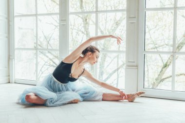 Genç Klasik Bale dansçısı kadın dans dersi. Mavi tutu etek beyaz ışık salonda büyük pencere yakınındaki güzel zarif balerin pratik bale pozisyonlar