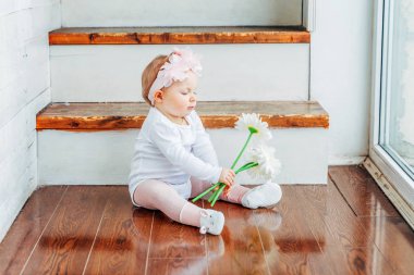 Küçük gülümseyen bebek kız bir yıl bahar kat pencere ve gerbera çiçekleri ile oynamaya yakın parlak ışık oturma odasında çelenk siting giymiş eski. Mutlu bir çocukluk evde oynamak. Çocukluk kavramı