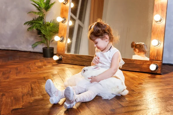 Маленькая девочка сидит на полу и играет с белым кроликом в современной комнате над зеркалом — стоковое фото