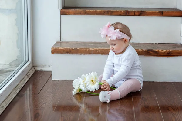 Kleine baby meisje dragen lente krans aanbrengen op de vloer in heldere lichte woonkamer in de buurt van venster en spelen met gerbera 's — Stockfoto